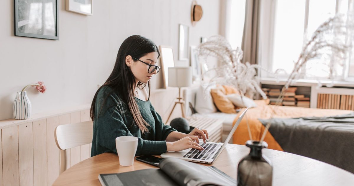 Side view of female remote worker in eyeglasses and casual wear typing on netbook while sitting at wooden table with cup and open magazine in bedroom at home