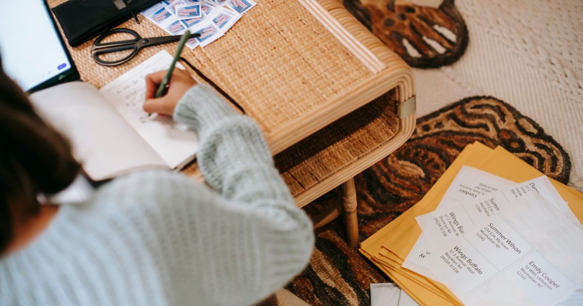 High angle of unrecognizable ethnic woman in casual outfit taking notes in notepad at table near laptop and scissors with stamps and shipping parcel bags on floor in light apartment