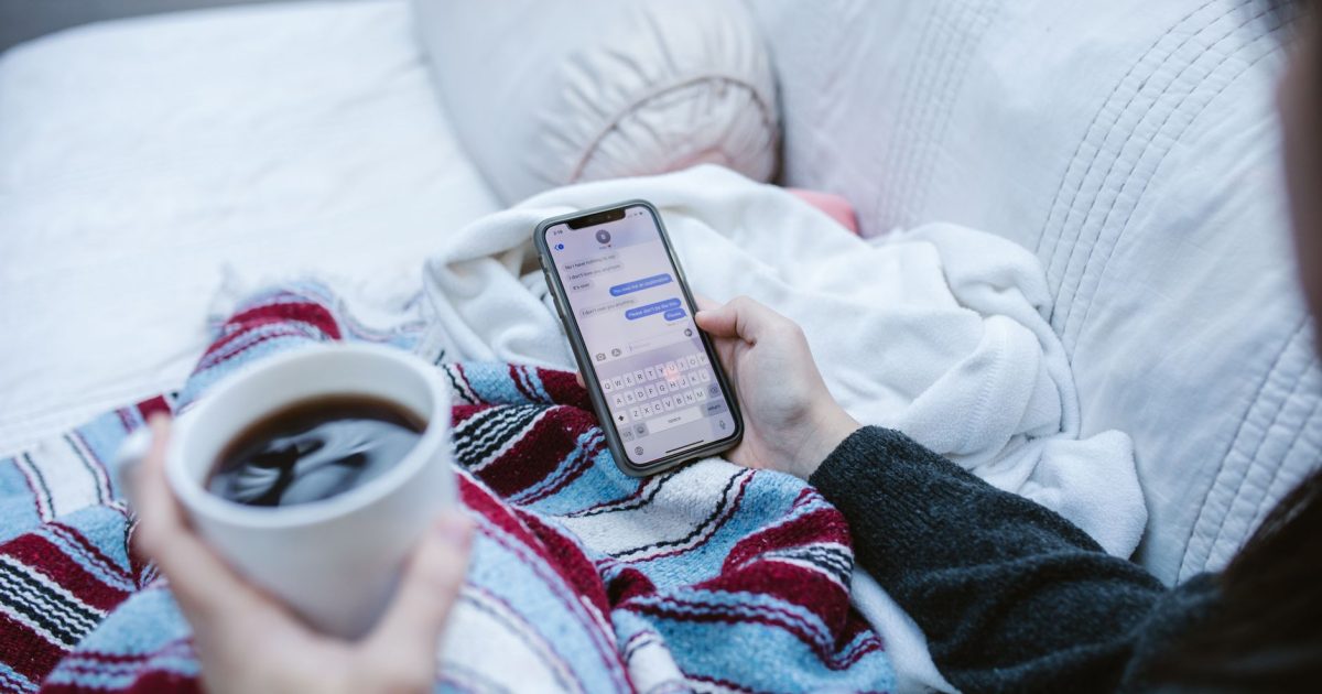 Person relaxing indoors with coffee and smartphone, texting in a cozy setting.