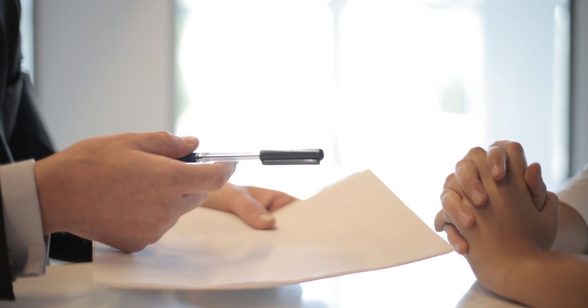 Side view of faceless formal man giving pen and paper to focused female with clenched hands at table on meeting