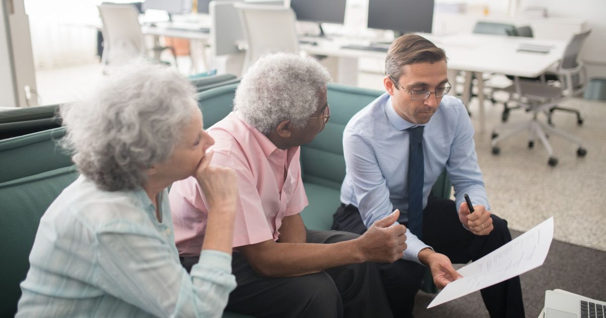 Business professional consults elderly clients in an office setting. Collaborative discussion, paperwork visible.
