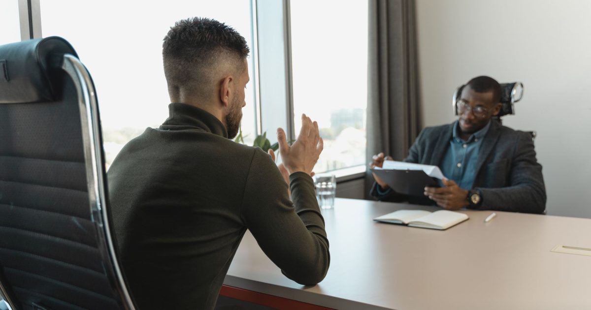 Two men in a job interview setting in a modern office with large windows.