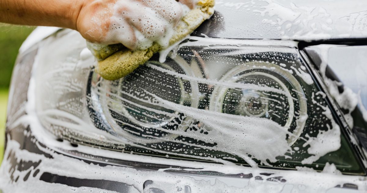 Hand scrubbing a car headlight with sponge and soap suds, detailing process.