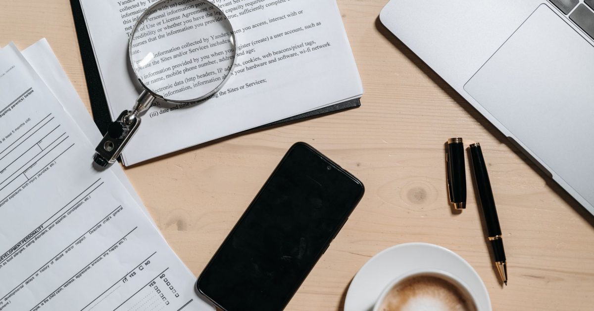 Top view of documents, laptop, coffee, and magnifying glass on office desk.