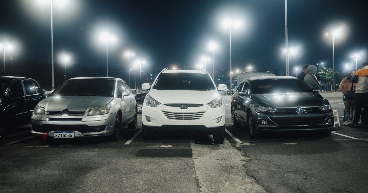 Night scene in a well-lit parking lot featuring various parked cars and street lamps.