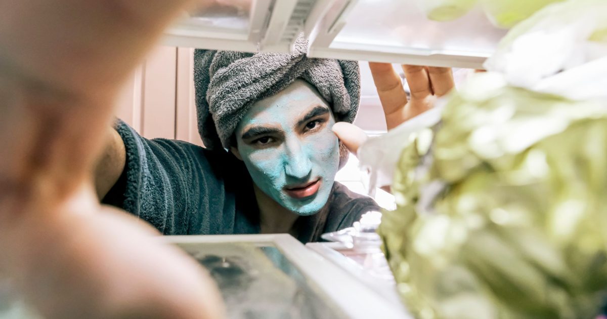 Person with a towel and face mask peering into an open fridge, surrounded by food.