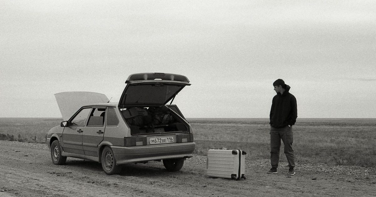 A man stands by a broken down car on a dirt road with luggage in a desolate, rural setting.