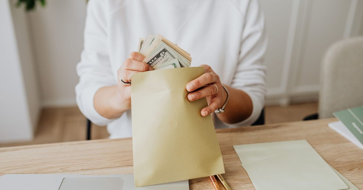 Close-up of person holding envelopes with cash at a wooden desk indoors.