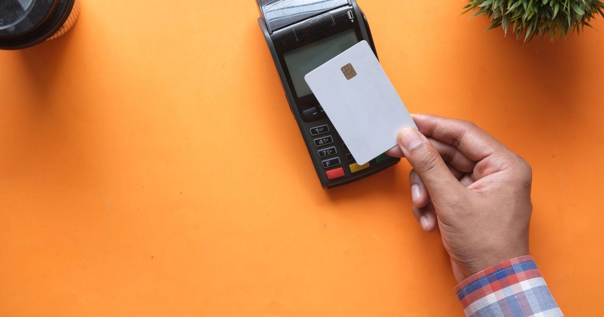 A person making a contactless payment with a credit card and card reader on a bright orange surface.